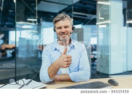 Experienced and successful businessman, looks at the webcam and smiles, talks to colleagues, online meeting, works in a modern office and uses a headset, video call 94335458