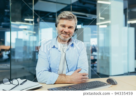 Experienced and successful businessman, looks at the webcam and smiles, talks to colleagues, online meeting, works in a modern office and uses a headset, video call Experienced and successful businessman, looks at the webcam and smiles, talks to colleagues, online meeting, works in a modern office and uses a headset, video call 94335459