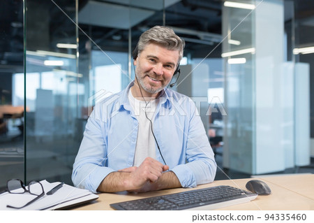Experienced and successful businessman, looks at the webcam and smiles, talks to colleagues, online meeting, works in a modern office and uses a headset, video call 94335460