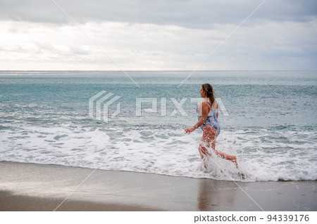 A plump woman in a bathing suit enters the water during the surf. Alone on the beach, Gray sky in the clouds, swimming in winter. A plump woman in a bathing suit enters the water during the surf. Alone on the beach, Gray sky in the clouds, swimming in winter. 94339176