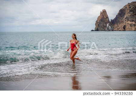 A beautiful and sexy brunette in a red swimsuit on a pebble beach, Running along the shore in the foam of the waves 94339280