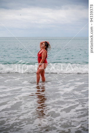 Woman in a bathing suit at the sea. A fat young woman in a red swimsuit enters the water during the surf 94339378