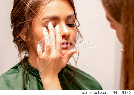 The hand of a makeup artist applies eye shadow on the eyelid of a young Caucasian woman with fingers in a beauty salon 94339380
