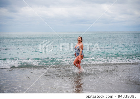 A plump woman in a bathing suit enters the water during the surf. Alone on the beach, Gray sky in the clouds, swimming in winter. 94339383