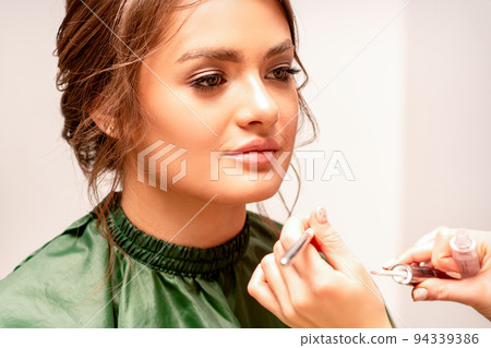 Makeup in the process. The hand of the makeup artist applies pink gloss lipstick on the lips of the beautiful face of the young caucasian woman in a beauty salon Makeup in the process. The hand of the makeup artist applies pink gloss lipstick on the lips of the beautiful face of the young caucasian woman in a beauty salon 94339386