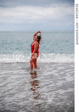 Woman in a bathing suit at the sea. A fat young woman in a red swimsuit enters the water during the surf 94339436