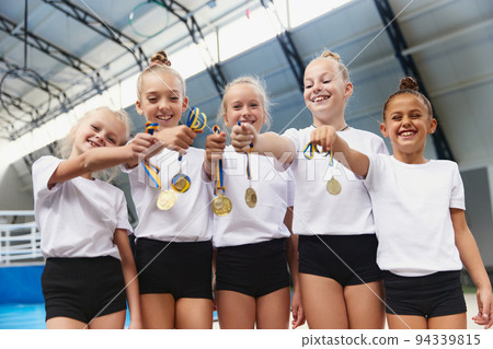 Happy little girls, female gymnastics athletes showing their medals at camera at sports gym, indoors. Concept of win, sport, studying, achievements and success Happy little girls, female gymnastics athletes showing their medals at camera at sports gym, indoors. Concept of win, sport, studying, achievements and success 94339815