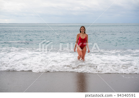 A beautiful and sexy brunette in a red swimsuit on a pebble beach, Running along the shore in the foam of the waves 94340591