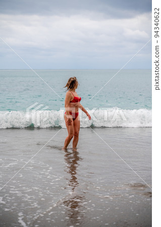 A middle-aged woman with a good figure in a red swimsuit on a pebble beach, running along the shore in the foam of the waves 94340622