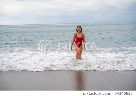 A beautiful and sexy brunette in a red swimsuit on a pebble beach, Running along the shore in the foam of the waves 94340623