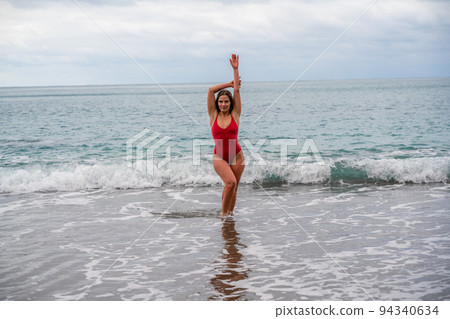 A beautiful and sexy brunette in a red swimsuit on a pebble beach, Running along the shore in the foam of the waves 94340634