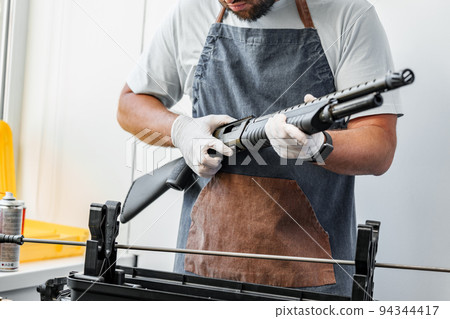 Close up of young man in apron disassembling a gun above the table Close up of young man in apron disassembling a gun above the table 94344417