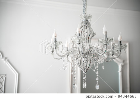 A beautiful expensive chandelier with many crystal crystals is hanging near a white wall. A beautiful expensive chandelier with many crystal crystals is hanging near a white wall. 94344663