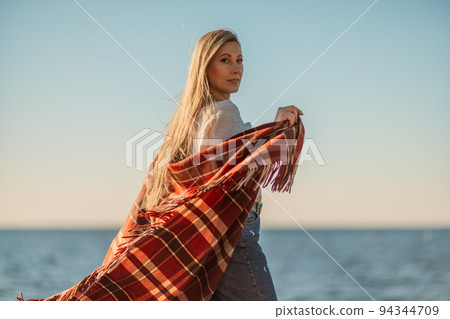 Attractive blonde Caucasian woman enjoying time on the beach at sunset, walking in a blanket and looking to the side, with the sunset sky and sea in the background. Beach vacation Attractive blonde Caucasian woman enjoying time on the beach at sunset, walking in a blanket and looking to the side, with the sunset sky and sea in the background. Beach vacation 94344709