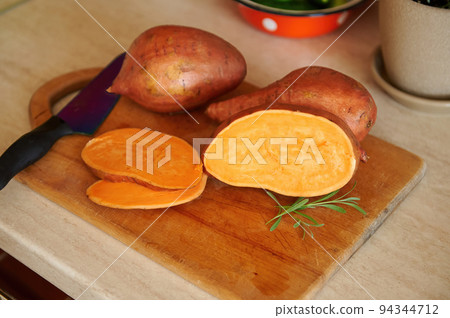 View from above of a whole and sliced tuber of organic raw orange color sweet potato, or batata, a kitchen knife and rosemary leaves for seasoning lying down on a wooden cutting board. 94344712