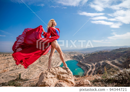 Side view of a beautiful sensual woman in a red long dress posing on a rock high above the lake in the afternoon. Against the background of the blue sky and the lake in the form of a heart Side view of a beautiful sensual woman in a red long dress posing on a rock high above the lake in the afternoon. Against the background of the blue sky and the lake in the form of a heart 94344721