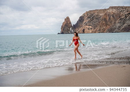 A beautiful and sexy brunette in a red swimsuit on a pebble beach, Running along the shore in the foam of the waves 94344735