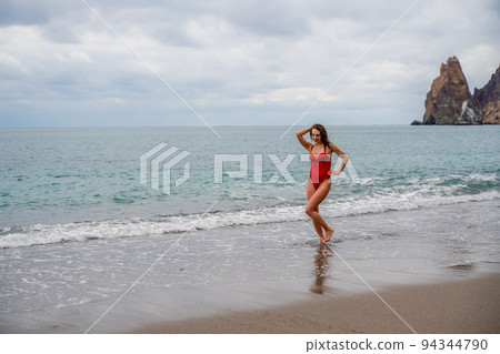 A beautiful and sexy brunette in a red swimsuit on a pebble beach, Running along the shore in the foam of the waves 94344790