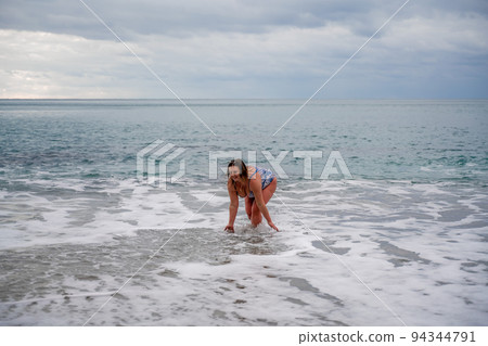 A plump woman in a bathing suit enters the water during the surf. Alone on the beach, Gray sky in the clouds, swimming in winter. A plump woman in a bathing suit enters the water during the surf. Alone on the beach, Gray sky in the clouds, swimming in winter. 94344791