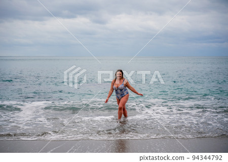 A plump woman in a bathing suit enters the water during the surf. Alone on the beach, Gray sky in the clouds, swimming in winter. A plump woman in a bathing suit enters the water during the surf. Alone on the beach, Gray sky in the clouds, swimming in winter. 94344792