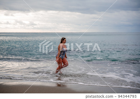 A plump woman in a bathing suit enters the water during the surf. Alone on the beach, Gray sky in the clouds, swimming in winter. A plump woman in a bathing suit enters the water during the surf. Alone on the beach, Gray sky in the clouds, swimming in winter. 94344793