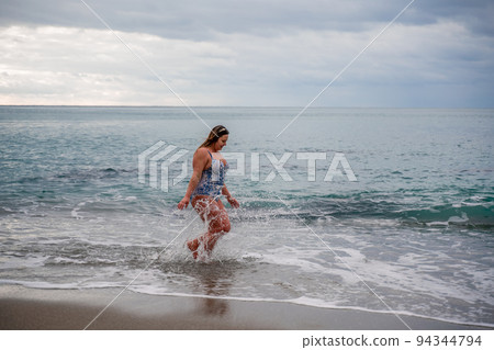 A plump woman in a bathing suit enters the water during the surf. Alone on the beach, Gray sky in the clouds, swimming in winter. A plump woman in a bathing suit enters the water during the surf. Alone on the beach, Gray sky in the clouds, swimming in winter. 94344794