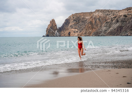 A beautiful and sexy brunette in a red swimsuit on a pebble beach, Running along the shore in the foam of the waves 94345316