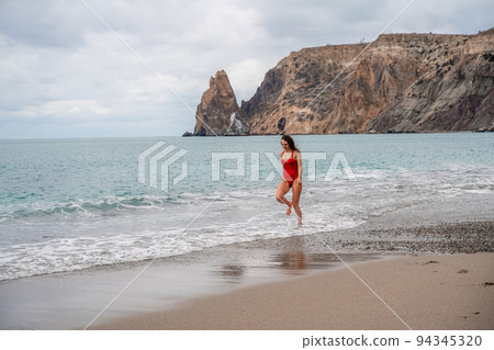 A beautiful and sexy brunette in a red swimsuit on a pebble beach, Running along the shore in the foam of the waves 94345320