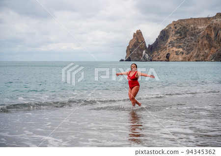 Woman in a bathing suit at the sea. A fat young woman in a red swimsuit enters the water during the surf 94345362
