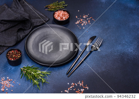 An empty black plate on a dark concrete background with spices and herbs An empty black plate on a dark concrete background with spices and herbs 94346232