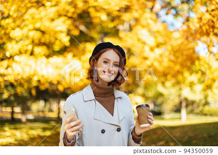 Glad caucasian millennial redhead lady in raincoat, beret chatting on smartphone, enjoy favorite drink in cup 94350059