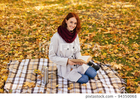 Glad caucasian millennial red-haired lady in raincoat with scarf reads book, sits on plaid in park with yellow leaves Glad caucasian millennial red-haired lady in raincoat with scarf reads book, sits on plaid in park with yellow leaves 94350068
