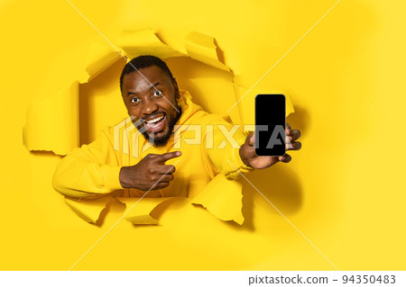 Excited black man showing cellphone with blank screen and pointing on it, posing through yellow paper torn hole 94350483