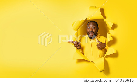Cheerful african american man pointing at free space and showing thumb up, looking through hole in yellow paper 94350616