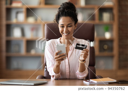 Positive businesswoman shopping online from office, using smartphone Positive businesswoman shopping online from office, using smartphone 94350762