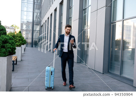 Happy confident handsome young caucasian male with beard in suit with suitcase goes, chatting by smartphone Happy confident handsome young caucasian male with beard in suit with suitcase goes, chatting by smartphone 94350904