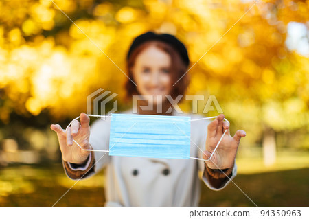 Glad european young red-haired woman in raincoat and beret puts on protective mask on camera in park 94350963