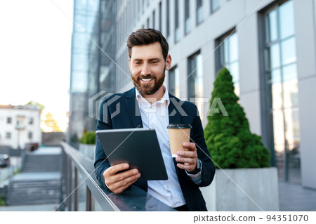 Smiling confident young caucasian guy with beard in suit has video call at tablet, hold cup of takeaway coffee 94351070
