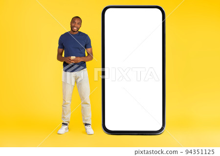African Guy Using Cellphone Standing Near Large Smartphone, Yellow Background 94351125