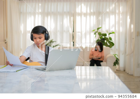 Asian boy playing guitar and watching online course on laptop while practicing for learning music or musical instrument online at home. Boy students study online with video call teachers play guitar. 94355684