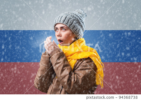 Energy crisis. Russian woman in hat, coat and scarf is warming herself. In the background is the flag of Russia and a snowstorm. Concept of sanctions, refusal of gas and renewable energy 94368738