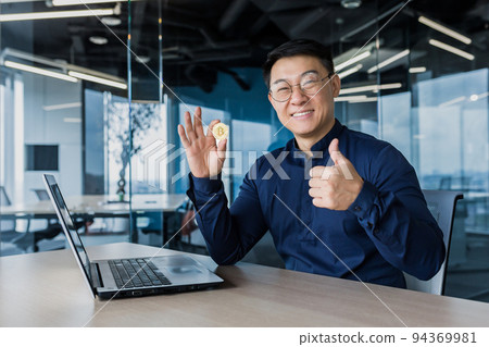 Successful asian businessman smiling and looking at camera, holding bitcoin coin and giving thumbs up, man working inside modern office building using laptop, broke stock exchange Successful asian businessman smiling and looking at camera, holding bitcoin coin and giving thumbs up, man working inside modern office building using laptop, broke stock exchange 94369981