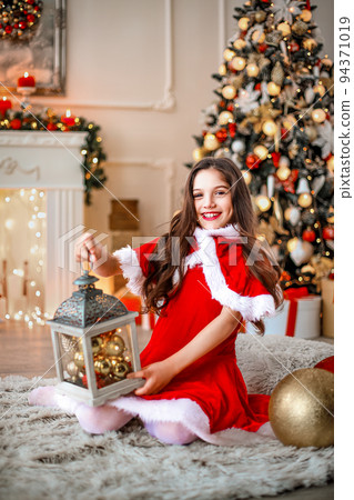 Cute little girl in checkered dress sitting in brightly decorated room and holding Christmas decoration 94371019