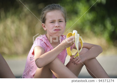 Young pretty child girl eating tasty ripe banana snacking outdoors on summer day Young pretty child girl eating tasty ripe banana snacking outdoors on summer day 94371274