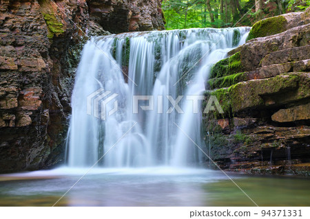 Waterfall on mountain river with white foamy water falling down from rocky formation in summer forest. Waterfall on mountain river with white foamy water falling down from rocky formation in summer forest. 94371331