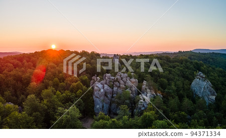Vibrant foggy evening over dark forest trees and large rocky formations at bright summer sunset. Amazingl scenery of wild woodland at dusk. 94371334