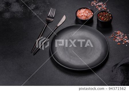 An empty black plate on a dark concrete background with spices, herbs and cutlery 94371562