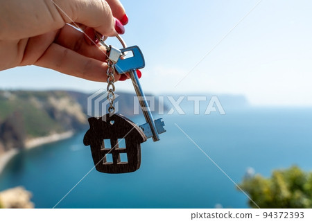 Keys with a keychain in the shape of a house in a female hand against the backdrop of the sea. The concept of buying a house, apartment, real estate, young family, mortgage loan, rental housing. Keys with a keychain in the shape of a house in a female hand against the backdrop of the sea. The concept of buying a house, apartment, real estate, young family, mortgage loan, rental housing. 94372393