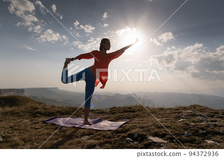 Woman doing yoga, Natarajasana asana - outdoor dance pose at sunset in the mountains. A yogi on a rug balances near a cliff during the day 94372936
