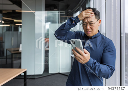 Asian businessman inside office building upset and sad reading bad news on mobile phone, business owner investor using smartphone standing near window 94373587